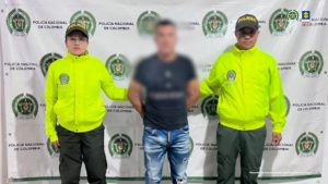 El capturado se encuentra de frente y con el rostro cubierto y custodiado por dos uniformados de la Policía Nacional. Detrás se observa un backing de la Policía Nacional.