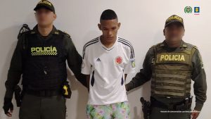 En la fotografía está un hombre de pie, joven, delgado, moreno, con los brazos atrás esposado, de cabellos negros crespos cortos. Se encuentra custodiado por dos uniformados de la Policía Nacional.