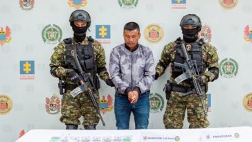 El capturado se encuentra de frente y en custodia por dos uniformados del Ejército Nacional. Detrás hay un backing del Ejército y en la foto se observa una mesa con el material que le incautaron.