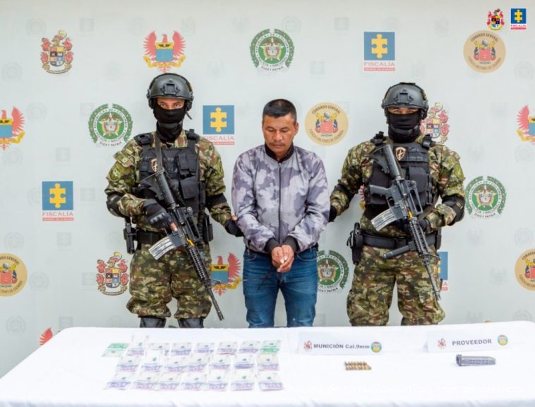 El capturado se encuentra de frente y en custodia por dos uniformados del Ejército Nacional. Detrás hay un backing del Ejército y en la foto se observa una mesa con el material que le incautaron.