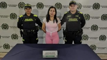 La mujer capturada se encuentra de frente y custodiada por dos uniformados de la Policía Nacional. Detrás hay un backing de la Policía.