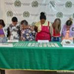 Los capturados se encuentran de frente y custodiados por dos uniformados de la Policía Nacional. Detrás hay un backing de la Policía Nacional. En la foto se observa una mesa con parte del material incautado como marihuana, dinero en efectivo y gramera.