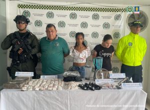 Se observan tres personas capturadas, en fila y de frente. A cada costado de la fila, un funcionario de la Policía Nacional. Atrás un pendón de Policía Nacional. Adelante una mesa en donde se observan las sustancias estupefacientes y otros elementos incautados en las diligencias de allanamiento y registro.