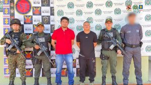 Los dos capturados se encuentran de frente y custodiados por un servidor del Cuerpo Técnico de Investigación CTI, dos uniformados del Gaula de la Policía y uno del Ejército. Detrás se observa un backing de la Policía y Ejército Nacional.