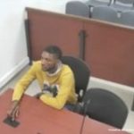 En la fotografía aparece un hombre delgado, moreno, con cabellos cortos, semi barba, vestido con un buzo amarillo, sentado en un escritorio de madera con un micrófono. Se encuentra al interior de la sala de audiencia donde se le procesó.