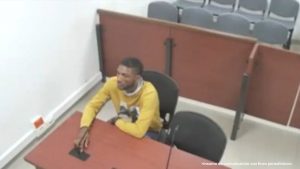 En la fotografía aparece un hombre delgado, moreno, con cabellos cortos, semi barba, vestido con un buzo amarillo, sentado en un escritorio de madera con un micrófono. Se encuentra al interior de la sala de audiencia donde se le procesó.