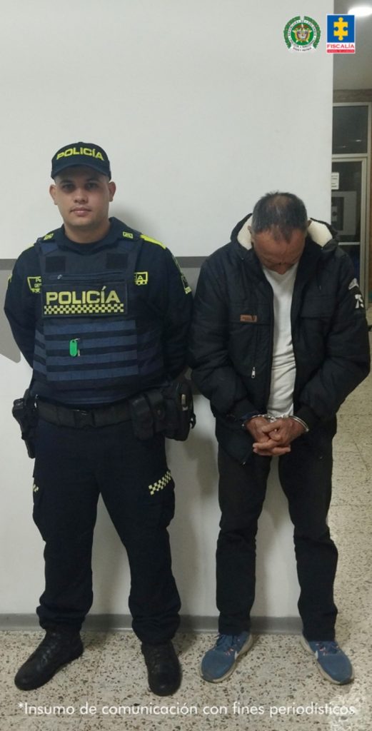 En la fotografía se aprecia de pie, con la cabeza agachada, los brazos adelante y esposado el adulto mayor. Viste chaqueta y pantalón de color negro, camiseta blanca y zapatillas azules. Al lado derecho de él hay un uniformado de la Policía Nacional.