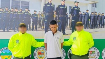 El capturado se encuentra de frente y custodiado por dos uniformados de la Policía Nacional. El capturado tiene un buzo blanco.