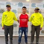 El capturado se encuentra de frente y custodiado por dos uniformados de la Policía Nacional. El capturado tiene una camiseta de color rojo y un jean