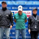 Fotografía del día de la captura del procesado usando una gorra que evita ver el rostro y dos servidores del CTI a los lados, en la parte de atrás hay un pendón azul con el logo de la Fiscalía