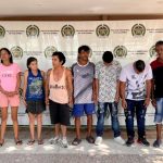 Los capturados se encuentran de frente y custodiados por dos uniformados de la Policía Nacional. Detrás se observa un backing de la Policía Nacional.