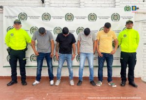 Los capturados se encuentran de frente  y  custodiados por dos uniformados de la Policía Nacional. Detrás se ve un backing de la Policía Nacional.