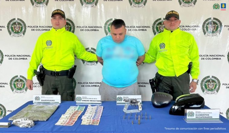 El capturado se encuentra de frente y custodiado por dos uniformados de la Policía Nacional. En la foto se observa el material incautado como marihuana, arma de fuego, munición, dinero en efectivo