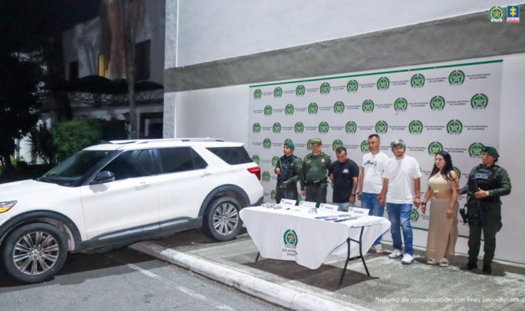 En la imagen se ven cuatro personas detenidas bajo custodia de integrantes de la Policía Nacional. Frente a ellos una mesa con varios elementos incautados. Detrás suyo un backing institucional.