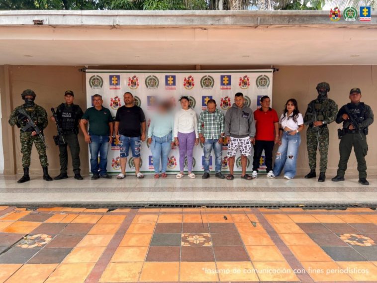 Los capturados se encuentran de frente y custodiados por dos uniformados del Gaula del Ejército y Policía Nacional. Detrás se observa un backing de la Policía Nacional con los logos de Ejército y Fiscalía.