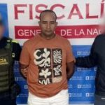 En la fotografía aparece un hombre de pie, moreno, con los brazos atrás esposado, la frente destapada, cabellos negros, crespos cortos. Se encuentra vestido con una camiseta café y custodiado por un uniformado del CTI y otro de la Policía Nacional.