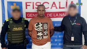 En la fotografía aparece un hombre de pie, moreno, con los brazos atrás esposado, la frente destapada, cabellos negros, crespos cortos. Se encuentra vestido con una camiseta café y custodiado por un uniformado del CTI y otro de la Policía Nacional.