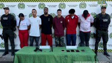 En la fotografía se aprecia en hilera y con los brazos atrás, a los seis judicializados, entre los que hay una mujer. A sus extremos hay uniformados del Gaula de la Policía Nacional. Delante de ellos hay una mesa con mantel verde oliva y sobre ella hay un arma de fuego, un proveedor y varios teléfonos celulares. Detrás de ellos hay un pendón institucional de la Policía.