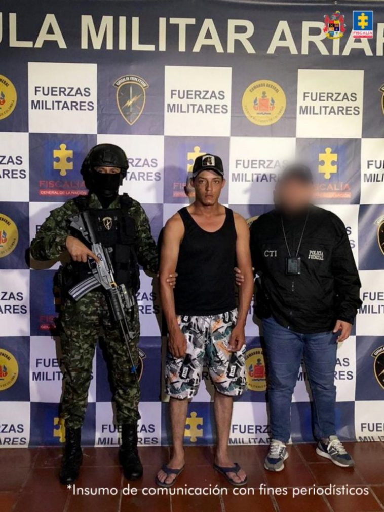 Capturado de frente custodiado por un investigador del CTI de la Fiscalía y un militar. Detrás banner del Grupo Gaula Ariari.