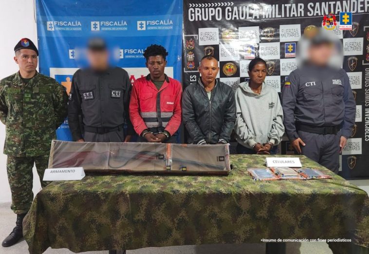 en la fotografia se puede ver a los dos detenidos en medios de dos funcionarios del CTI y un militar, atras de ellos se encuentra un baking de la Fiscalia y el Ejercito. en frente de ellos esta una mesa en donde exhiben un arma y celulares incautados