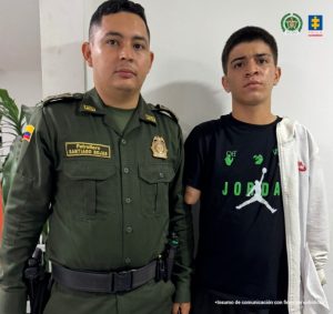 En la fotografía aparece Darío Johan Montoya Valencia de pie. Lleva una camiseta negra de manga corta; a la altura del pecho impreso con la palabra “Jordan” y números verdes; Bermudas negras y busto blanco. A su derecha está un miembro uniformado de la policía nacional.