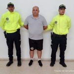 En la fotografía está un hombre de pie con los brazos atrás, de tez blanca, contextura gruesa, calvo, vestido con una camiseta gris, una bermuda y tenis negros. Se encuentra custodiado por dos uniformados de la Policía Nacional.