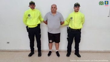 En la fotografía está un hombre de pie con los brazos atrás, de tez blanca, contextura gruesa, calvo, vestido con una camiseta gris, una bermuda y tenis negros. Se encuentra custodiado por dos uniformados de la Policía Nacional.