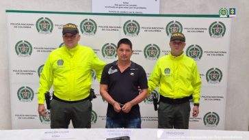 El capturado se encuentra de frente y  custodiado por dos uniformados de la Policía Nacional. En la foto se ve de fondo un backing de la Policía Nacional.