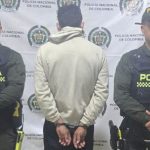 El capturado se encuentra de espalda y custodiado por dos uniformados de la Policía Nacional. En la foto se ve de fondo un backing de la Policía Nacional.