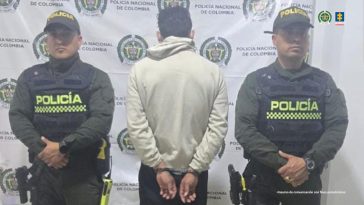 El capturado se encuentra de espalda y custodiado por dos uniformados de la Policía Nacional. En la foto se ve de fondo un backing de la Policía Nacional.