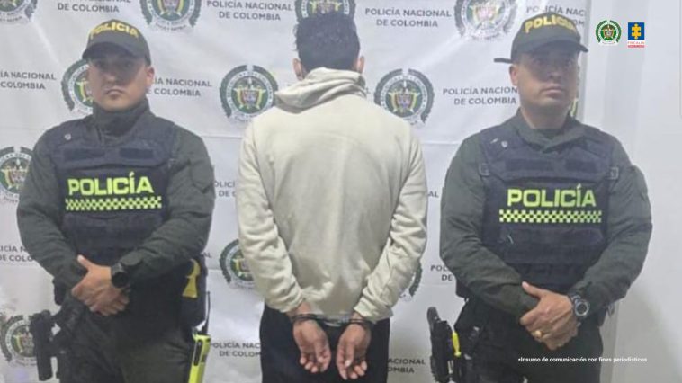 El capturado se encuentra de espalda y custodiado por dos uniformados de la Policía Nacional. En la foto se ve de fondo un backing de la Policía Nacional.