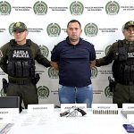 El capturado aparece con sus manos esposadas a la espalda, y se encuentra custodiado por uniformados del Gaula de la Policía Nacional. Detrás de ellos un pendón institucional.