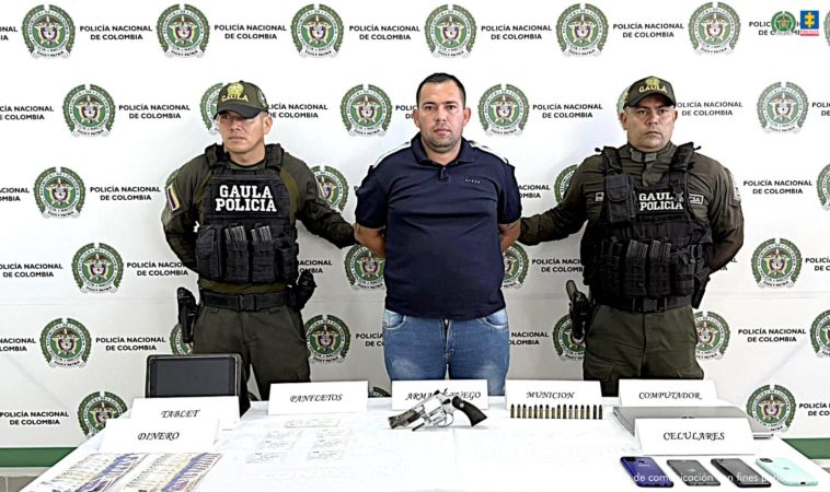 El capturado aparece con sus manos esposadas a la espalda, y se encuentra custodiado por uniformados del Gaula de la Policía Nacional. Detrás de ellos un pendón institucional.