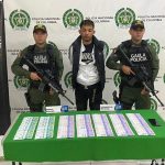 El capturado aparece detrás de una mesa en la que hay billetes de diferentes denominaciones. Dos personas del Gaula rodean al capturado. Al fondo se ve un banner de la Policía.