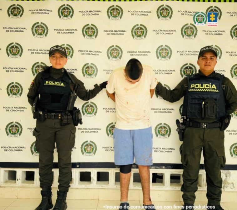 El capturado se encuentra de frente y custodiado por dos uniformados de la Policía Nacional. En la foto se ve de fondo un backing de la Policía Nacional.