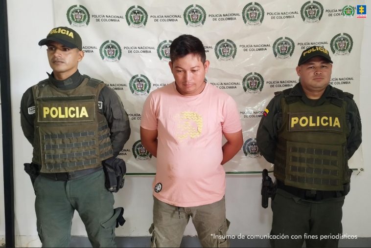 El capturado aparece en el centro de la imagen, con las manos esposadas en la espalda, rodeado de dos miembros de la Policía. Al fondo puede verse un banner de la Policía.