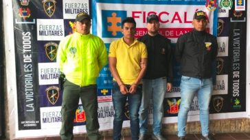 El capturado se encuentra de frente y custodiado por servidores del Cuerpo Técnico de Investigación CTI, uniformado de la Policía Nacional y Gaula. En la foto se ve de fondo un backing de la Fiscalía, Policía y Ejército Nacional.