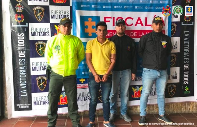 El capturado se encuentra de frente y custodiado por servidores del Cuerpo Técnico de Investigación CTI, uniformado de la Policía Nacional y Gaula. En la foto se ve de fondo un backing de la Fiscalía, Policía y Ejército Nacional.