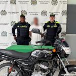 En la imagen aparece un hombre de suéter blanco, entre dos uniformados de la Policía y frente a una mesa y una motocicleta. 