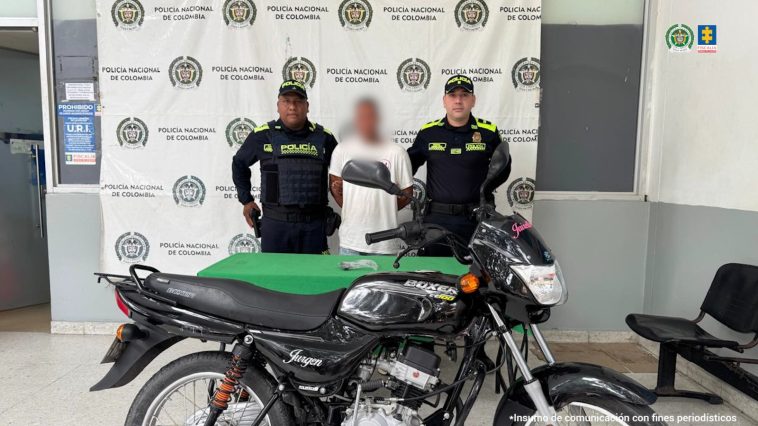 En la imagen aparece un hombre de suéter blanco, entre dos uniformados de la Policía y frente a una mesa y una motocicleta. 