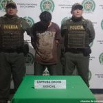 En la fotografía aparece un hombre de pie, con los brazos atrás esposado, con la cabeza agachada, moreno, de pelo negro, vestido con un camiseta café y un jean negro. Se encuentra custodiado por dos uniformados de la Policía Nacional. 