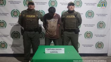 En la fotografía aparece un hombre de pie, con los brazos atrás esposado, con la cabeza agachada, moreno, de pelo negro, vestido con un camiseta café y un jean negro. Se encuentra custodiado por dos uniformados de la Policía Nacional. 