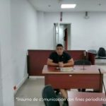 En la fotografía está un hombre de tez trigueña, cabellos negros, vestido con camiseta negrar, sentado en un escritorio de madera con un micrófono en el interior de la sala de audiencia donde se le procesó. 