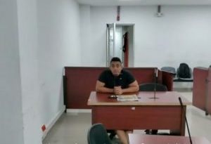 En la fotografía está un hombre de tez trigueña, cabellos negros, vestido con camiseta negrar, sentado en un escritorio de madera con un micrófono en el interior de la sala de audiencia donde se le procesó. 