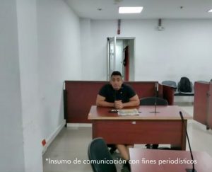 En la fotografía está un hombre de tez trigueña, cabellos negros, vestido con camiseta negrar, sentado en un escritorio de madera con un micrófono en el interior de la sala de audiencia donde se le procesó. 