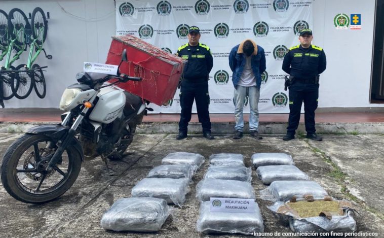 El capturado se encuentra de frente y custodiado por dos uniformados de la Policía Nacional. En la foto se ve de fondo un backing de la Policía Nacional. En la foto se observa una motocicleta y el estupefaciente incautado.