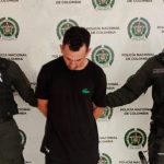 El capturado se encuentra de frente con la cabeza agachada y custodiado por dos uniformados de la Policía Nacional. En la foto se ve de fondo un backing de la Policía Nacional.