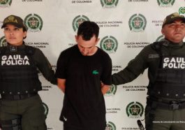 El capturado se encuentra de frente con la cabeza agachada y custodiado por dos uniformados de la Policía Nacional. En la foto se ve de fondo un backing de la Policía Nacional.