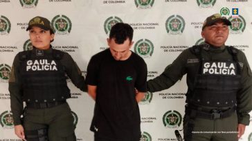 El capturado se encuentra de frente con la cabeza agachada y custodiado por dos uniformados de la Policía Nacional. En la foto se ve de fondo un backing de la Policía Nacional.