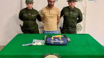 El capturado aparece detrás de una mesa, en la que están los elementos incautados. A cada lado suyo hay miembros de la Policía.